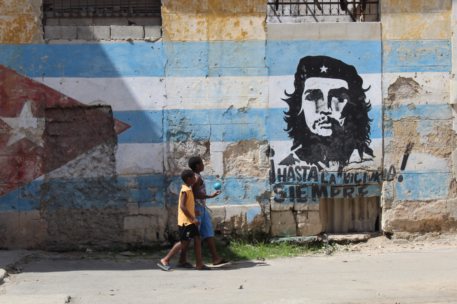 Two children walking past a weathered Cuban flag and Che Guevara mural in Havana